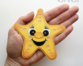 Puede incluir: Una estrella de mar amarilla con una cara sonriente, ojos negros y una nariz naranja. La estrella de mar mide 4,25 pulgadas por 4,25 pulgadas.