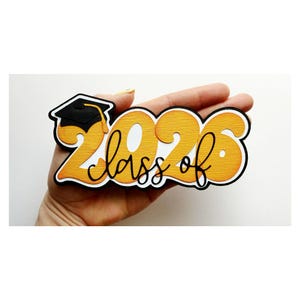 May include: A graduation-themed cutout with the text "Class of 2026" in black script over large orange numbers. A small black graduation cap with a gold tassel is in the upper left corner. The cutout is held in a hand.