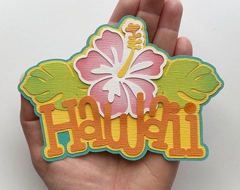 May include: A paper craft with the word "Hawaii" in orange and yellow letters with a teal outline. The letters are layered and have a textured appearance. A pink hibiscus flower with a white center and yellow stamen is above the word. Green leaves with a textured appearance are on either side of the flower.