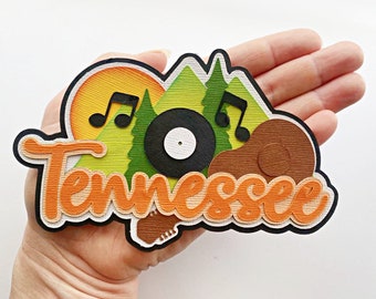 May include: A paper craft design featuring the word "Tennessee" in orange and white lettering with a layered paper design. The design also includes a green mountain scene with a black vinyl record, a brown guitar, and musical notes.