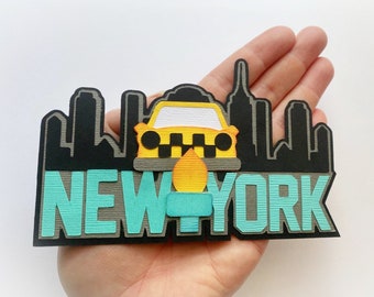 May include: A paper craft depicting the New York City skyline with a yellow taxi cab in the center. The skyline is black with gray accents and the word "NEW YORK" is written in teal and gray letters.