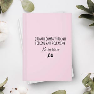 May include: A pink journal with the text "GROWTH COMES THROUGH FEELING AND RELEASING" and the name "Katarina" in a script font. A black heart-shaped hand symbol is below the name. The journal is on top of a white journal.