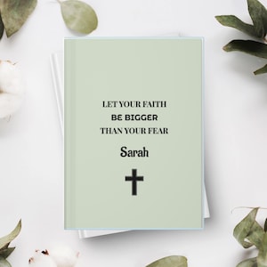 Puede incluir: Un cuaderno de color verde salvia con el texto en negro "LET YOUR FAITH BE BIGGER THAN YOUR FEAR" seguido del nombre "Sarah" en una fuente estilizada y un símbolo de cruz negra simple centrado en la portada del diario rectangular.
