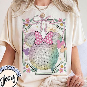 May include: Ivory t-shirt with a Minnie Mouse-themed graphic. The design includes two geometric spheres, a pink polka-dot bow, and Mickey Mouse shapes. The shirt also features the words "COMFORT COLORS" and "Ivory". The design is set against a floral and ribbon background.