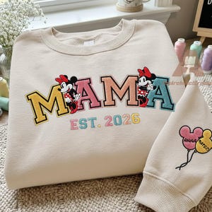 May include: Cream-colored sweatshirt with the word "MAMA" embroidered in colorful letters, featuring Minnie Mouse figures. The text "EST. 2026" is below. A matching sleeve with Mickey Mouse balloons is also present.