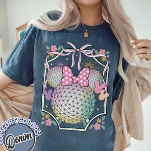 May include: Navy blue t-shirt with a graphic of a Minnie Mouse bow and ears, set against a geometric sphere design. The design includes pink, yellow, and blue hues, with floral accents and fireworks. The text "COMFORT COLORS Denim" is on the bottom left.