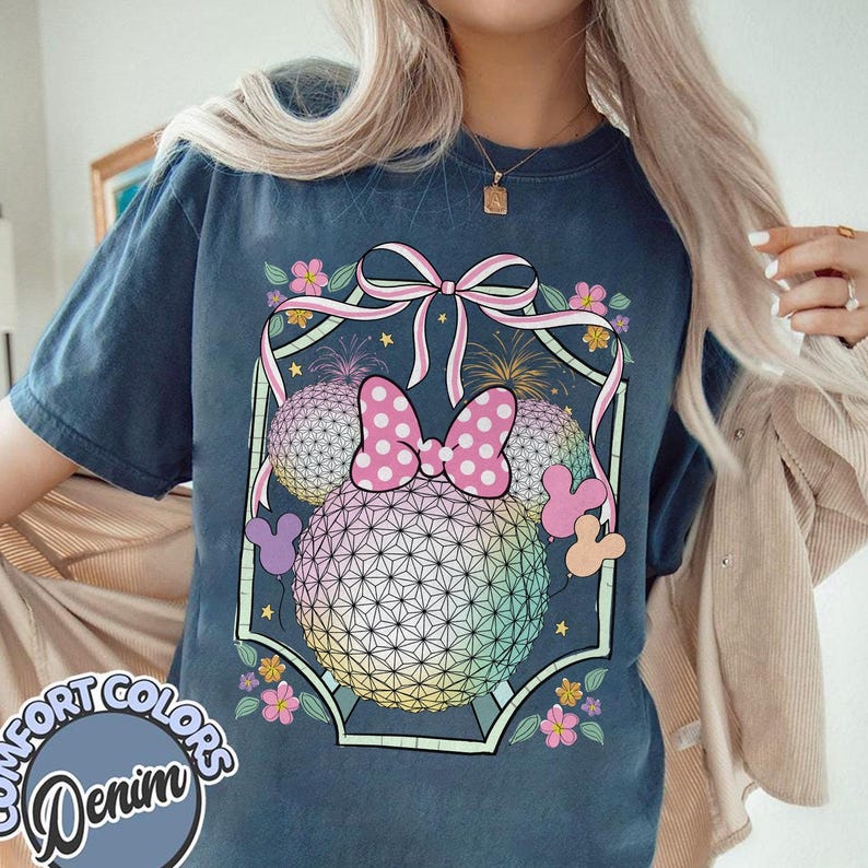 May include: Navy blue t-shirt with a graphic of a Minnie Mouse bow and ears, set against a geometric sphere design. The design includes pink, yellow, and blue hues, with floral accents and fireworks. The text "COMFORT COLORS Denim" is on the bottom left.