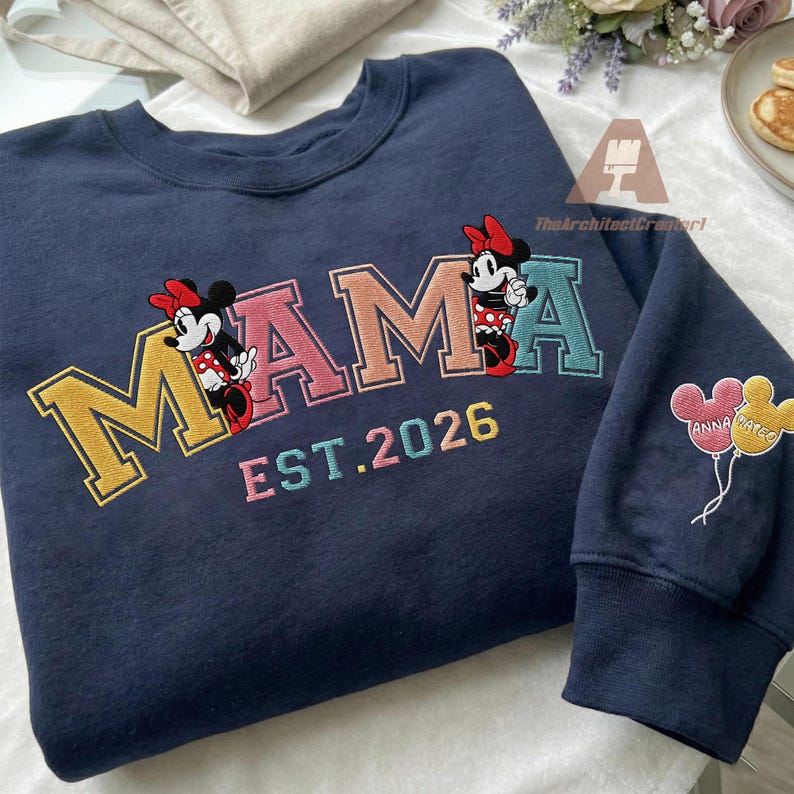 May include: Navy blue sweatshirt with the word "MAMA" in colorful embroidered letters, featuring Minnie Mouse characters. The text "EST. 2026" is below the word "MAMA". The sleeve has balloons with names.