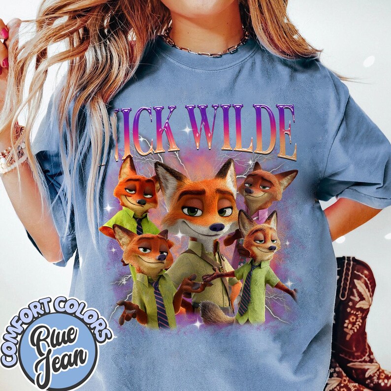Puede incluir: Camiseta azul claro Comfort Colors con un gr&aacute;fico de Nick Wilde de Zootopia. El dise&ntilde;o incluye el texto "Nick Wilde" en una fuente degradada e im&aacute;genes de Nick Wilde en varias poses. Tambi&eacute;n es visible un logotipo circular con las palabras "Comfort Colors" y "Blue Jean".