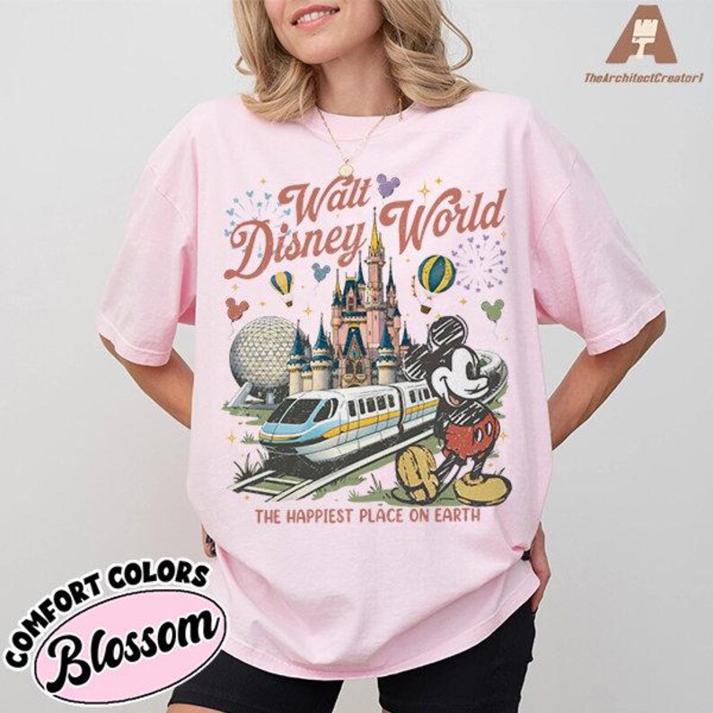 May include: Pink t-shirt with a graphic of Walt Disney World, featuring a castle, train, and Mickey Mouse. The text "The Happiest Place on Earth" is below the graphic. The shirt also has the text "Comfort Colors Blossom".