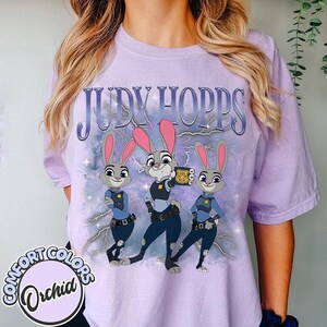 May include: A light purple t-shirt featuring the text "JUDY HOPPS" above a graphic of three cartoon bunnies in police uniforms. The shirt has a Comfort Colors label with the word "Orchid" in a circle.