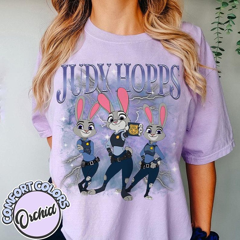 May include: A light purple t-shirt featuring the text "JUDY HOPPS" above a graphic of three cartoon bunnies in police uniforms. The shirt has a Comfort Colors label with the word "Orchid" in a circle.