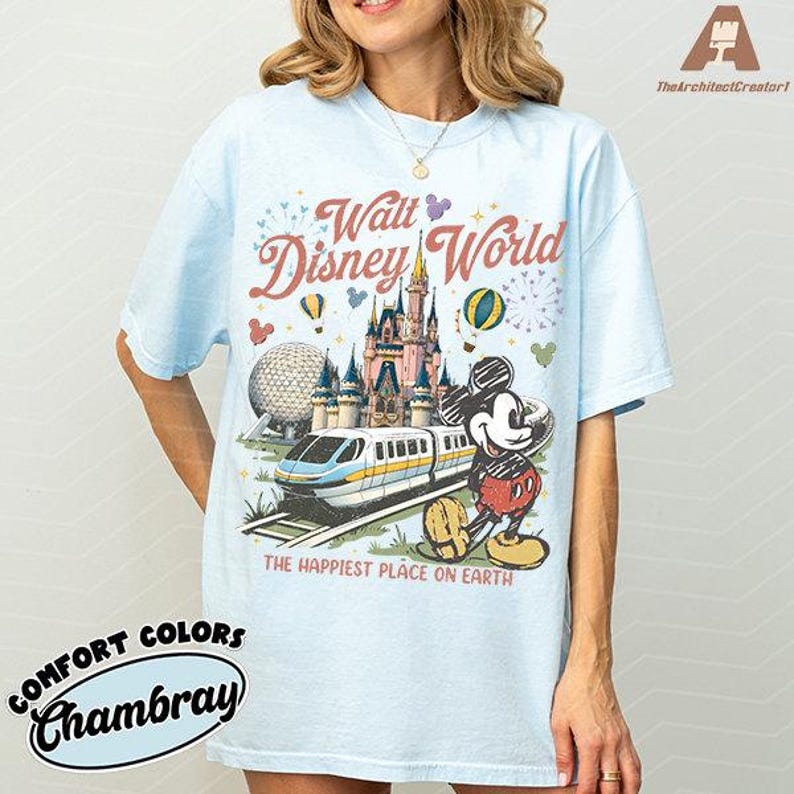 May include: Light blue t-shirt featuring a colorful Walt Disney World graphic. The design includes a castle, Mickey Mouse, a monorail, and the text "The Happiest Place on Earth". The shirt is made of Comfort Colors Chambray fabric.