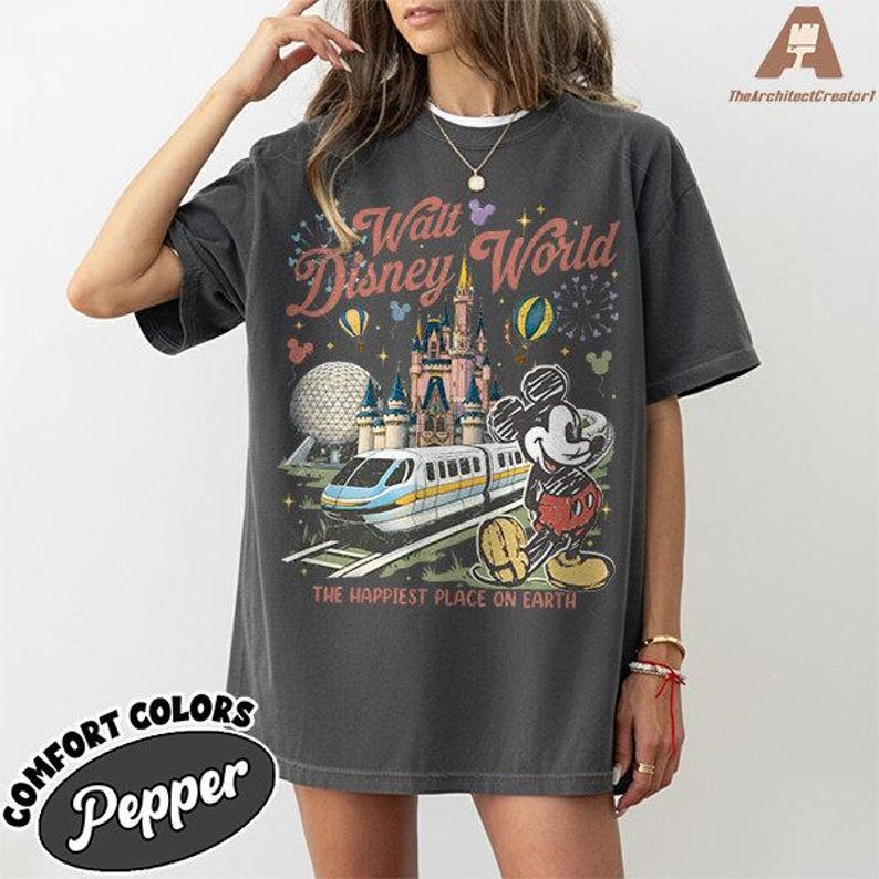 May include: Dark grey t-shirt featuring a colorful graphic of Walt Disney World. The design includes Cinderella's castle, Mickey Mouse, a monorail, and the text "The Happiest Place on Earth". The shirt is labeled "Comfort Colors Pepper".
