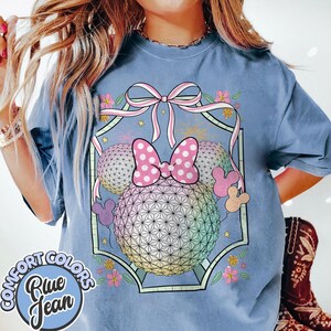 May include: Blue jean Comfort Colors t-shirt with a graphic of a geometric sphere and a pink polka dot bow, surrounded by flowers and Mickey Mouse ears. The shirt is light blue and has a round Comfort Colors logo.