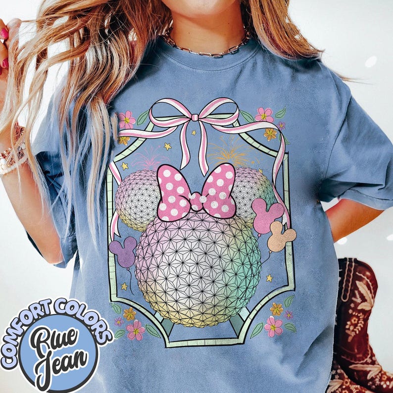 May include: Blue jean Comfort Colors t-shirt with a graphic of a geometric sphere and a pink polka dot bow, surrounded by flowers and Mickey Mouse ears. The shirt is light blue and has a round Comfort Colors logo.