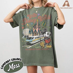 May include: Moss green t-shirt featuring a colorful graphic of Walt Disney World with Mickey Mouse. The design includes a castle, monorail, and the text "World Disney World" and "The Happiest Place on Earth". The shirt also has the text "Comfort Colors" and "Moss".