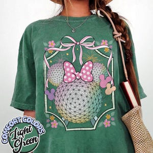 May include: Light green Comfort Colors t-shirt with a Minnie Mouse-themed graphic. The design includes a pink polka-dot bow, two geometric spheres, and fireworks. The shirt has the words "Comfort Colors" and "Light Green" printed on it.