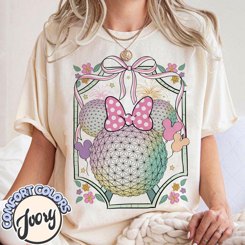 May include: Ivory t-shirt with a Minnie Mouse-themed graphic. The design includes two geometric spheres, a pink polka-dot bow, and Mickey Mouse shapes. The shirt also features the words "COMFORT COLORS" and "Ivory". The design is set against a floral and ribbon background.