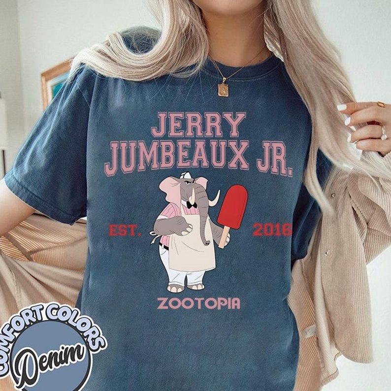 Puede incluir: Una camiseta azul vaquera con el texto "JERRY JUMBEAUX JR." sobre una ilustraci&oacute;n de un elefante sosteniendo un helado rojo. La camiseta tambi&eacute;n incluye "EST. 2018" y "ZOOTOPIA" en rosa.