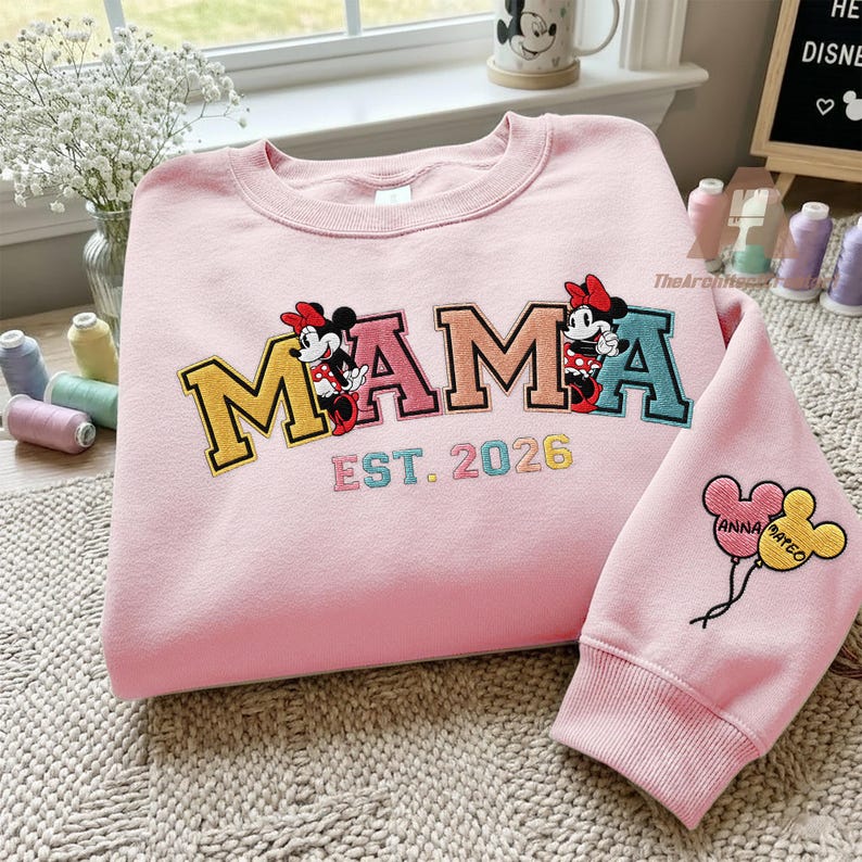 May include: A light pink sweatshirt with the word "MAMA" embroidered in yellow, pink, and teal, featuring Minnie Mouse characters. The text "EST. 2026" is below. A matching sleeve has balloon graphics with names.