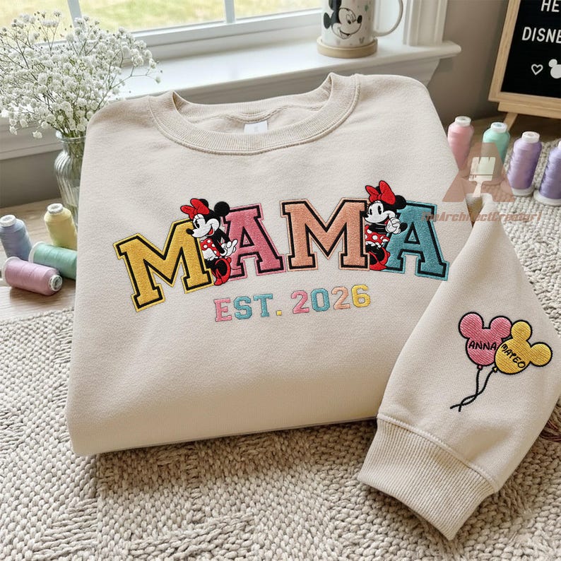 May include: Cream-colored sweatshirt with the word "MAMA" embroidered in colorful letters, featuring Minnie Mouse figures. The text "EST. 2026" is below. A matching sleeve with Mickey Mouse balloons is also present.