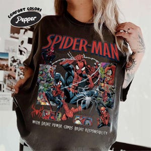 K&ouml;nnte beinhalten: Dunkelgraues T-Shirt mit einem Spider-Man-Motiv. Das Design zeigt die roten Worte "SPIDER-MAN" und Comic-Panels mit Spider-Man in Aktion. Der Text "WITH GREAT POWER COMES GREAT RESPONSIBILITY" ist ebenfalls enthalten.