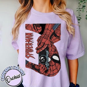 Puede incluir: Camiseta Comfort Colors color orqu&iacute;dea con un gr&aacute;fico desgastado de Spider-Man. El dise&ntilde;o incluye la palabra "SPIDER-MAN" en rojo, una c&aacute;mara vintage y las manos y m&aacute;scara ic&oacute;nicas del personaje. La camiseta es de color morado claro.