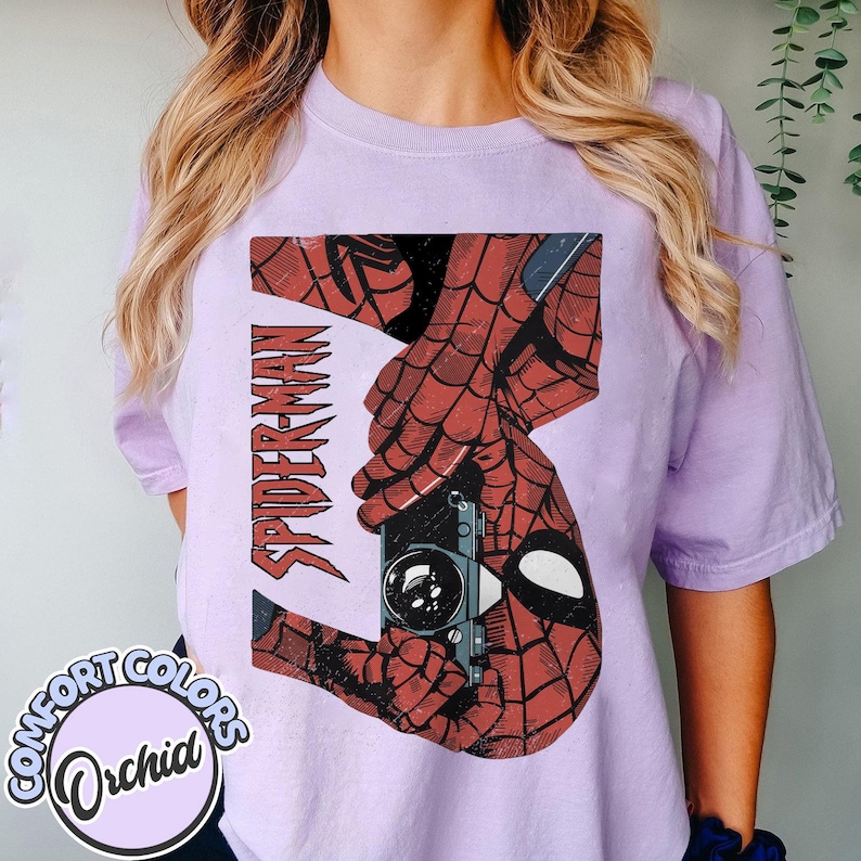 Puede incluir: Camiseta Comfort Colors color orqu&iacute;dea con un gr&aacute;fico desgastado de Spider-Man. El dise&ntilde;o incluye la palabra "SPIDER-MAN" en rojo, una c&aacute;mara vintage y las manos y m&aacute;scara ic&oacute;nicas del personaje. La camiseta es de color morado claro.