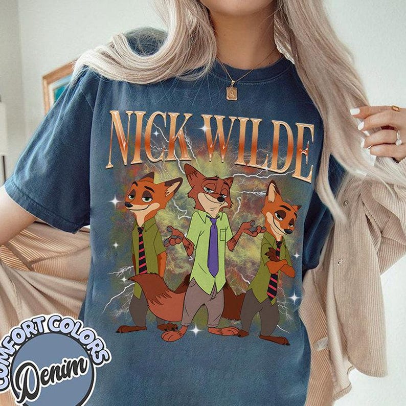 May include: A navy blue t-shirt featuring the text "NICK WILDE" above a graphic of three anthropomorphic foxes. The foxes are wearing shirts and ties, with lightning bolts in the background. The shirt has a "Comfort Colors Denim" logo.