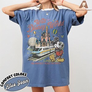 May include: Blue jean-colored t-shirt featuring a graphic of Walt Disney World. The design includes a castle, monorail, Mickey Mouse, and the text "Walt Disney World" and "The Happiest Place on Earth". The shirt also has the text "Comfort Colors".