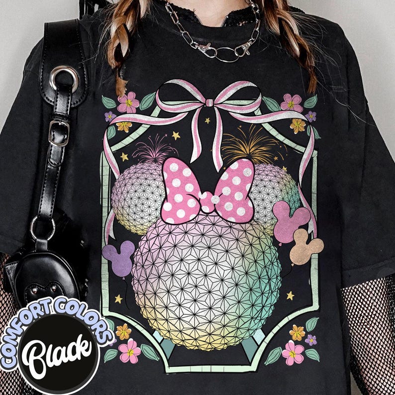 May include: Black Comfort Colors t-shirt with a colourful graphic design. The design features two spherical shapes with a geometric pattern, a pink polka-dot bow, and floral and fireworks elements. The shirt has a distressed appearance.