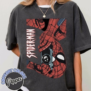 Puede incluir: Camiseta gris oscuro con un gr&aacute;fico de Spider-Man rojo y negro. El dise&ntilde;o incluye la palabra "SPIDER-MAN" en blanco, y una imagen estilizada de las manos de Spider-Man sosteniendo una c&aacute;mara. La camiseta tiene un logo de Comfort Colors y Pepper.