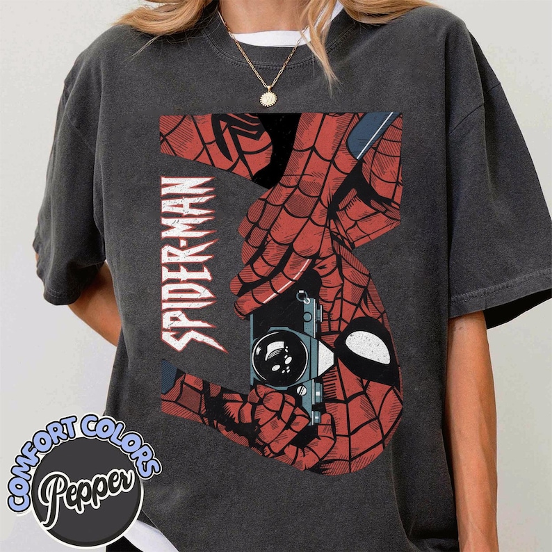 Puede incluir: Camiseta gris oscuro con un gr&aacute;fico de Spider-Man rojo y negro. El dise&ntilde;o incluye la palabra "SPIDER-MAN" en blanco, y una imagen estilizada de las manos de Spider-Man sosteniendo una c&aacute;mara. La camiseta tiene un logo de Comfort Colors y Pepper.