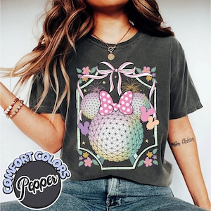 May include: Dark grey t-shirt with a colourful graphic of Minnie Mouse's head, bow, and geometric spheres. The design includes fireworks, flowers, and a pink bow. The shirt has the words "COMFORT COLORS Pepper" in a circle.