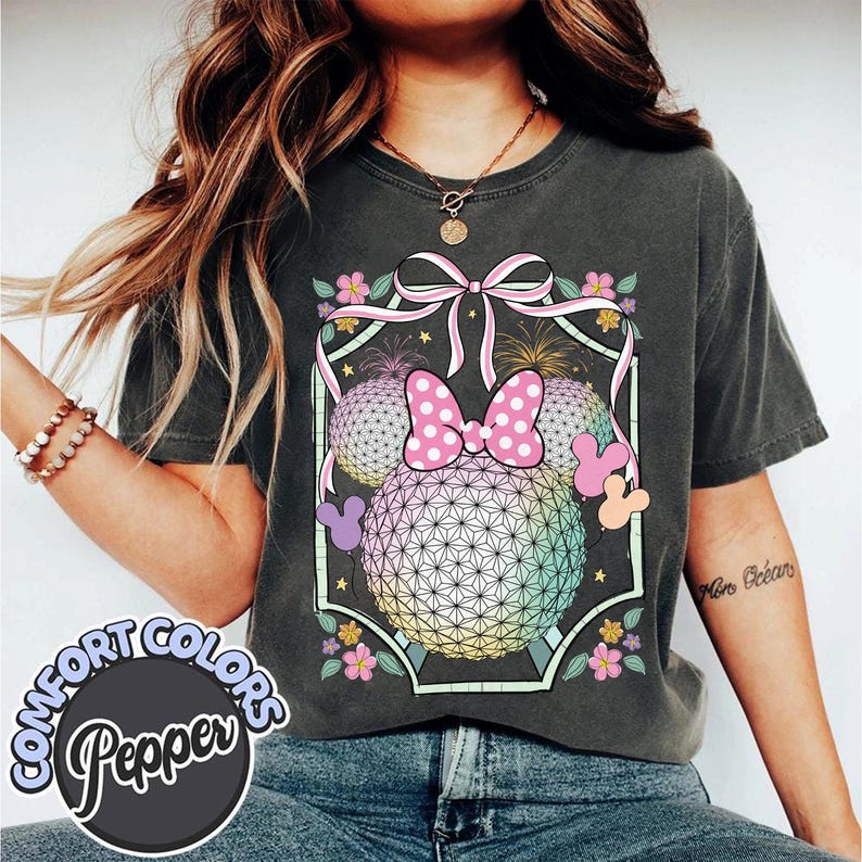 May include: Dark grey t-shirt with a colourful graphic of Minnie Mouse's head, bow, and geometric spheres. The design includes fireworks, flowers, and a pink bow. The shirt has the words "COMFORT COLORS Pepper" in a circle.