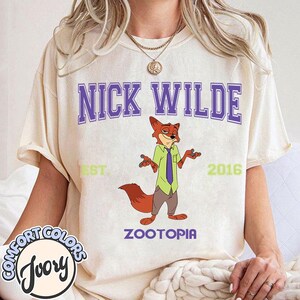 Puede incluir: Camiseta color crema con la inscripci&oacute;n "NICK WILDE" en letras moradas en bloque sobre un personaje de zorro de dibujos animados. El zorro lleva una camisa verde y una corbata morada. La palabra "ZOOTOPIA" est&aacute; debajo.