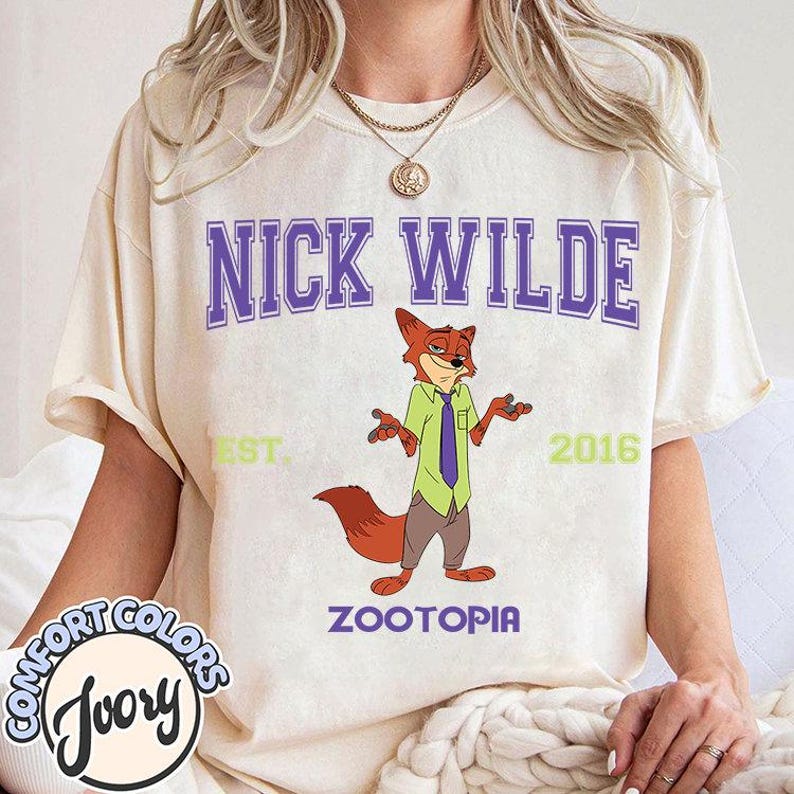 Puede incluir: Camiseta color crema con la inscripci&oacute;n "NICK WILDE" en letras moradas en bloque sobre un personaje de zorro de dibujos animados. El zorro lleva una camisa verde y una corbata morada. La palabra "ZOOTOPIA" est&aacute; debajo.
