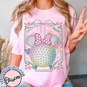 May include: Pale pink t-shirt with a graphic design featuring a bow, two spheres, and Mickey Mouse ears. The spheres have a geometric pattern and a rainbow gradient. The shirt also has the text "Comfort Colors Blossom" in a circle.