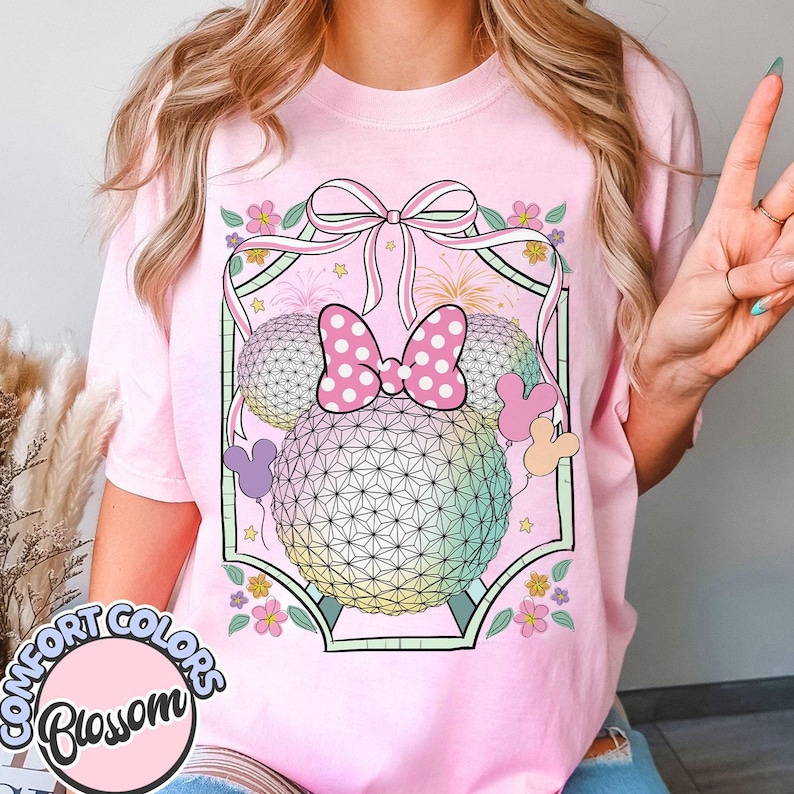 May include: Pale pink t-shirt with a graphic design featuring a bow, two spheres, and Mickey Mouse ears. The spheres have a geometric pattern and a rainbow gradient. The shirt also has the text "Comfort Colors Blossom" in a circle.