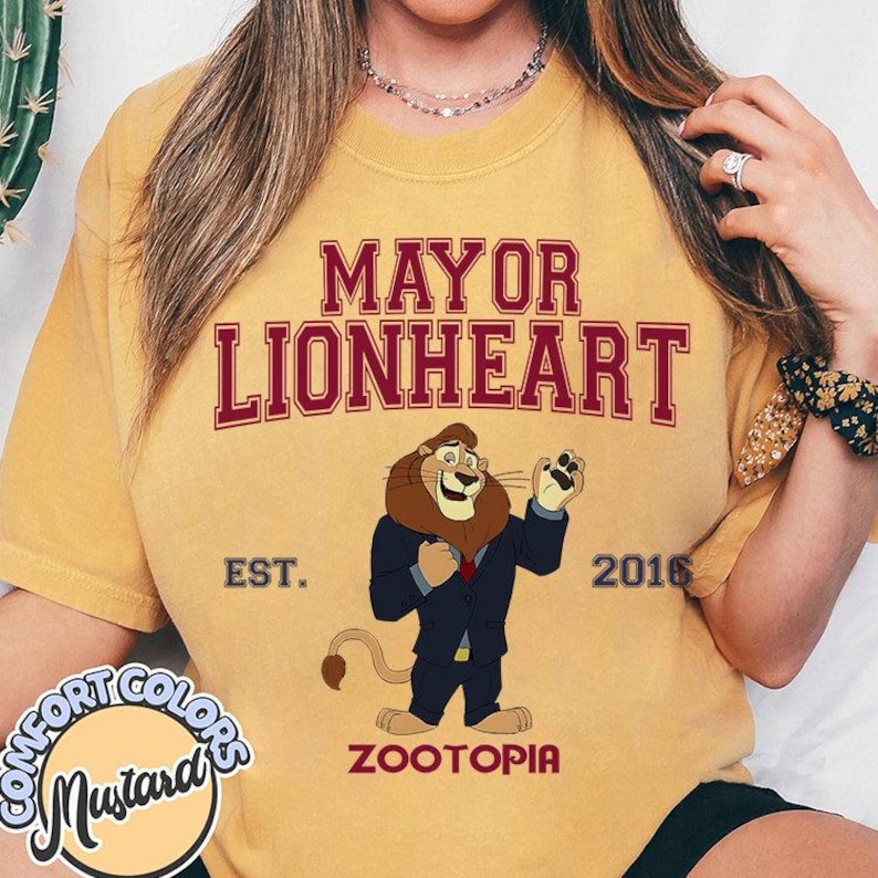 Puede incluir: Camiseta amarilla mostaza con el texto "MAYOR LIONHEART" y una ilustraci&oacute;n de un le&oacute;n con traje. La camiseta tambi&eacute;n incluye el texto "EST. 2016" y "ZOOTOPIA".