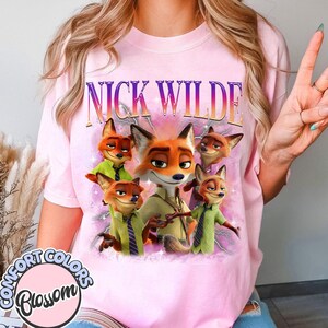 Puede incluir: Camiseta rosa claro con un dise&ntilde;o de Nick Wilde de Zootr&oacute;polis. El dise&ntilde;o incluye m&uacute;ltiples im&aacute;genes del personaje zorro, el texto "NICK WILDE" en degradado y el logotipo Comfort Colors Blossom.