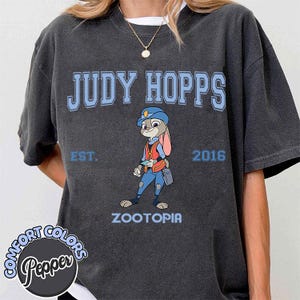 Puede incluir: Camiseta gris oscuro con el texto "JUDY HOPPS" en azul claro, y una ilustraci&oacute;n de Judy Hopps de Zootopia. La camiseta tambi&eacute;n incluye "EST. 2016" y "ZOOTOPIA". La camiseta es de Comfort Colors.