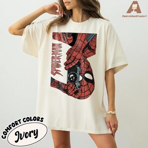 Puede incluir: Camiseta color marfil con un gr&aacute;fico desgastado de Spider-Man sosteniendo una c&aacute;mara. El dise&ntilde;o incluye el texto "SPIDER-MAN" en fuente vertical. La camiseta es parte de la colecci&oacute;n "Comfort Colors".