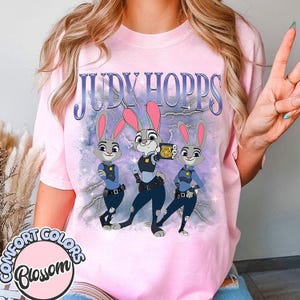 May include: Pink t-shirt featuring the text "JUDY HOPPS" above a graphic of three cartoon bunnies in police uniforms. The bunnies are gray with pink ears and are set against a purple and blue background with lightning bolts. The shirt has the "COMFORT COLORS Blossom" logo.