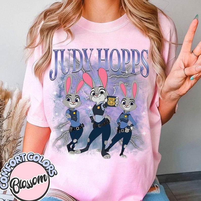 May include: Pink t-shirt featuring the text "JUDY HOPPS" above a graphic of three cartoon bunnies in police uniforms. The bunnies are gray with pink ears and are set against a purple and blue background with lightning bolts. The shirt has the "COMFORT COLORS Blossom" logo.
