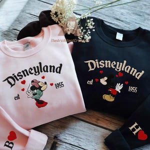 Op de afbeelding: Twee sweatshirts, één roze en één zwart, met Disneyland-borduurwerk. Het roze sweatshirt heeft Minnie Mouse, terwijl de zwarte Mickey Mouse heeft. Beide hebben de tekst "Disneyland est 1955" en rode hartaccenten.