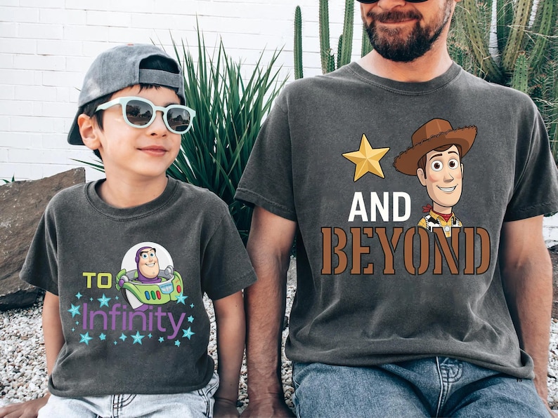 May include: Two charcoal gray t-shirts with graphics from the movie Toy Story. One shirt features Buzz Lightyear with the text "To Infinity" and the other features Woody with the text "And Beyond".