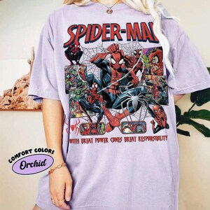 K&ouml;nnte beinhalten: Orchideenfarbenes T-Shirt mit einem Spider-Man-Motiv. Das Design enth&auml;lt die roten Worte "SPIDER-MAN" und verschiedene Spider-Man-Illustrationen. Der Text "WITH GREAT POWER COMES GREAT RESPONSIBILITY" ist ebenfalls sichtbar.