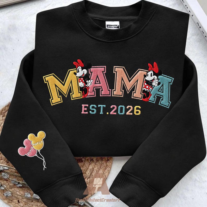 May include: Black sweatshirt with the word "MAMA" embroidered in yellow, pink, and teal, featuring Minnie Mouse characters. The text "EST.2026" is below the word "MAMA". The left sleeve has embroidered balloons with the names "Anna" and "Kate".